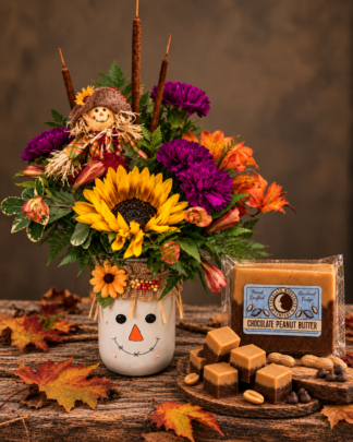 Scarecrow’s Sweet Harvest fall flower arrangement featuring golden sunflower, purple carnations, orange blooms, smiling scarecrow keepsake vase, and gourmet chocolate peanut butter fudge from Aggieland Flowers & Chocolates.